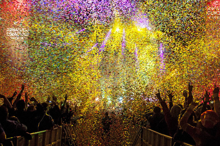 Fotografia colorida com papeis picados, na maioria em roxo e amarelo, caindo sobre o público durante um show