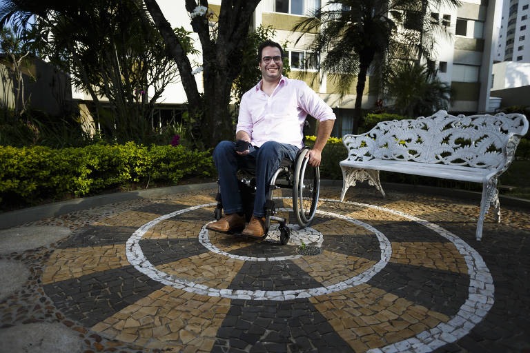 Bruno Mahfuz, de 37 anos, fundador do guiaderodas, posa para foto no térreo de seu prédio, em São Paulo; ele está sentado em sua cadeira de rodas e veste uma camisa branca, calças jeans e usa óculos. Está numa área aberta, arborizada, e há um banco branco atrás dele