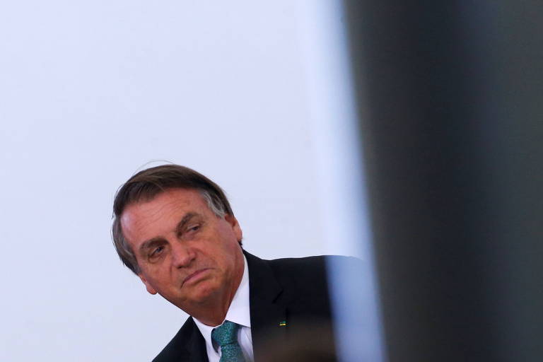 Bolsonaro, um homem branco, com cabelo liso, castanho e grisalho nas pontas, fazendo uma expressão de descontentamento. Ele está de terno e aparece na altura do peito, atrás de uma parede