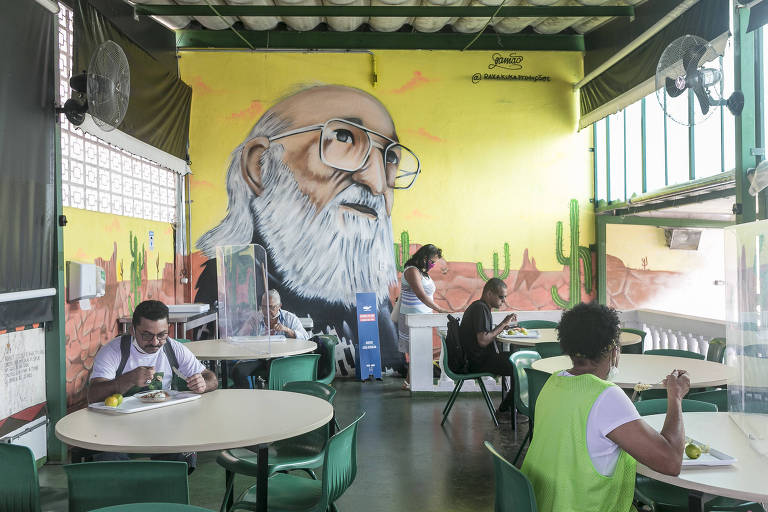 Imagem mostra o refeitório do Cieja, escola na zona sul de São Paulo. Há cinco pessoas na foto, três mulheres e dois homens. Quatro dessas pessoas estão sentadas, fazendo suas refeições, enquanto uma das mulheres está deixando o ambiente. Numa das paredes do refeitório, há uma pintura com o rosto do educador Paulo Freire, em fundo amarelo