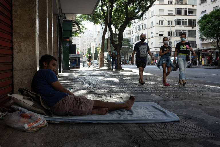 Jovem em situação de rua está deitado debaixo de uma marquise enquanto pedestres passam e olham para ele 