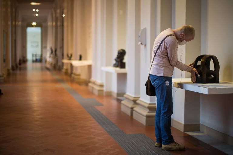 Um homem está em pé e usando máscara em um corredor longo; ele está com o corpo levemente encurvado para a frente e toca as mãos em uma escultura de metal