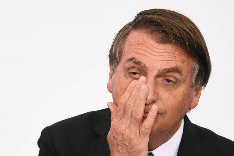 Bolsonaro aparece dos ombros para cima, esfregando a mão direita sobre o olho.