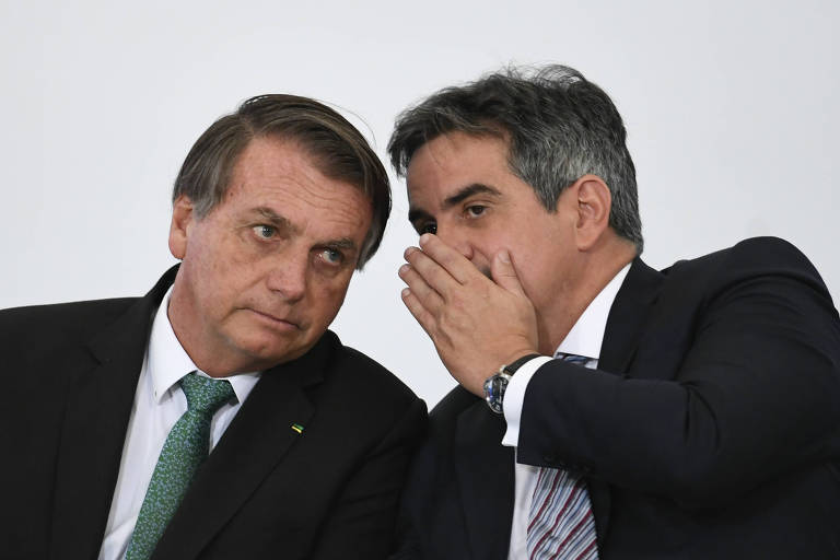 Dois homens de terno escuro e gravata sentados lado a lado conversando. Um deles cobre a boca para falar com o outro, que escuta com atenção.