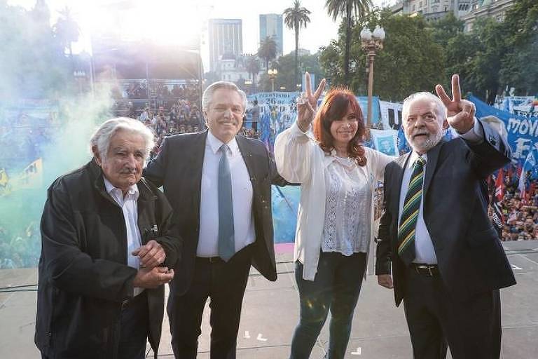 Lula, Critina Kirchner, Alberto Fernández e Pepe Mujica em palco, com multidão segurando bandeiras e lançando fumaça ao fundo