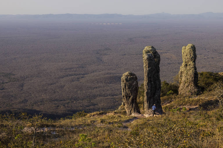 Três pedras muito altas se destacam no alto de um morro. Uma pessoa passa perto delas, parecendo ser minúscula, na comparação. Ao fundo há uma vale, com vegetação rasa.