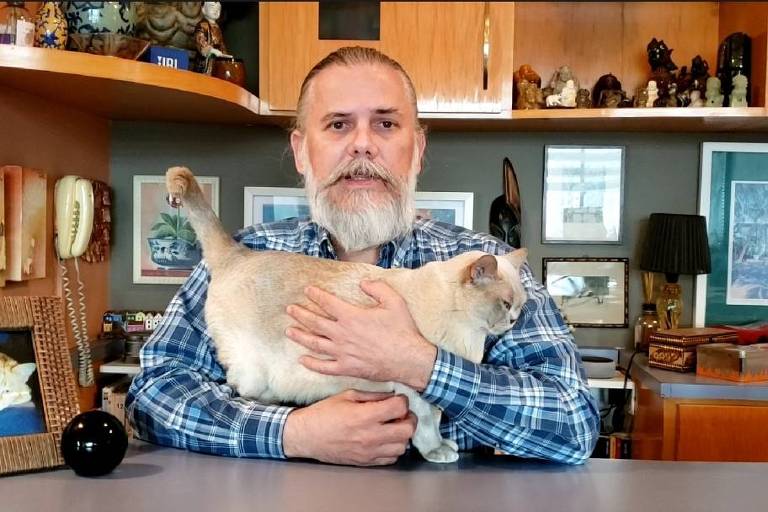 Gerson Alves Pereira é um homem branco de cabelos e barba grisalhos; ele veste uma camisa xadrez azul e segura uma gata da raça burmês de cor creme