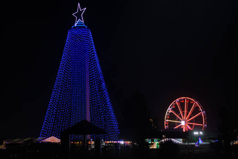 Foto noturna mostra árvore de Natal feita de luzes azuis ao lado de roda-gigante com iluminação vermelha