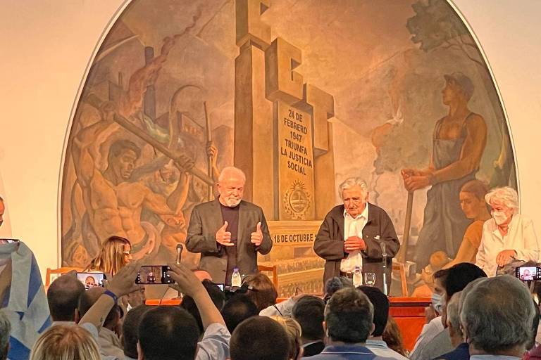 Dois homens estão em um palco tendo um mural pintado na parede atrás de si; à esquerda está Lula e à direita, José Mujica; ambos são observados por uma plateia sentada  