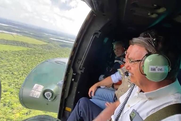 Imagem feita dentro helicóptero mostra um homem de meia idade, o presidente Jair Bolsonaro, sorrindo e com fone, olhando através da porta da aeronave as áreas atingidas por chuvas na Bahia