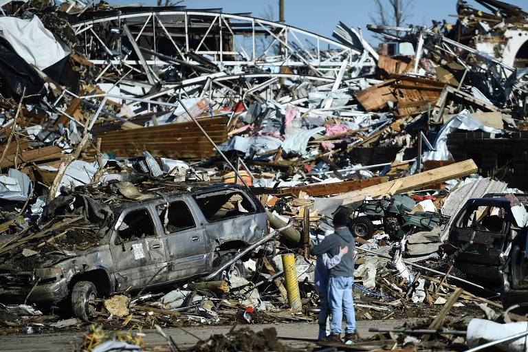 Duas pessoas abraçadas observam uma massa de escombros empilhadas, incluindo vigas e uma caminhonete; a cena retrata os estragos provocados por tornados em Mayfield, Kentucky