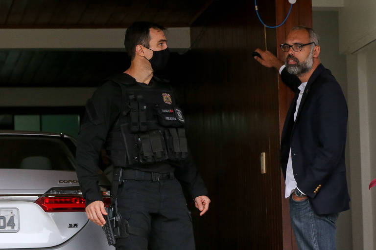 Homem de óculos conversa com policial federal de máscara na garagem de sua casa