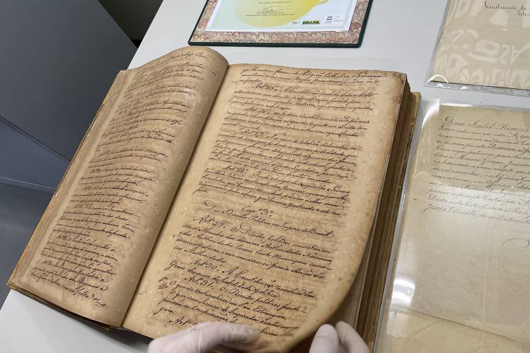 Livro antigo, com páginas manuscritas e amareladas pelo tempo, aberto sobre uma mesa. Veem-se as mãos de um funcionário, com  luvas, manuseando as páginas do volume.