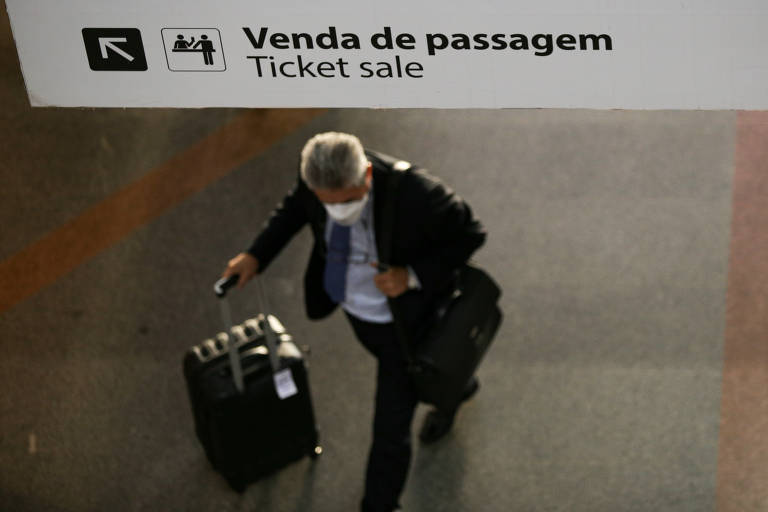 Imagem apérea mostra homem de máscara arrastando uma mala com rodinhas em corredor de um aeroporto.