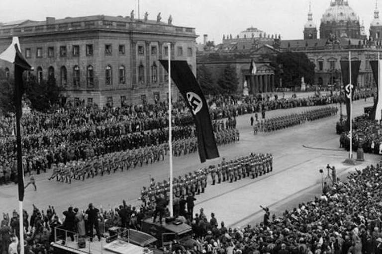 Imagem aérea mostra um aglomerado de pessoas assistindo a um desfile. Ao centro, outro grupo de pessoas marcham. Bandeiras com o símbolo nazista estão hasteadas