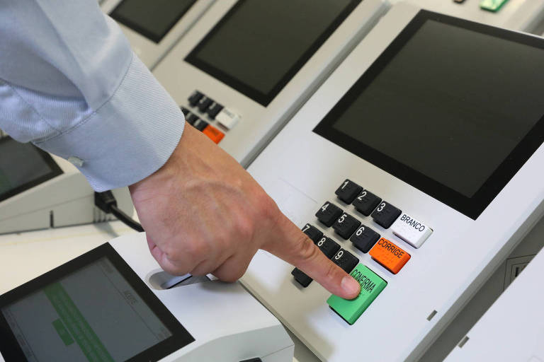 Mão de pessoa branca apertando botão confirma verde na urna eletrônica