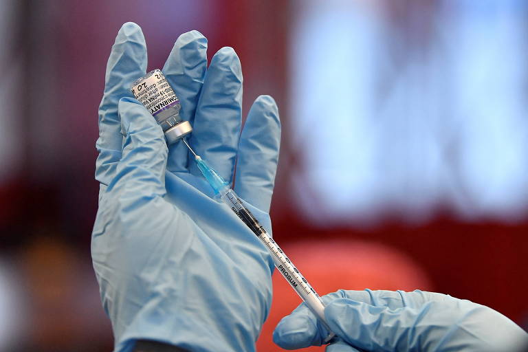 Imagem mostra as duas mãos de mulher com luvas, uma delas segurando a seringa e a outra, a ampola com a vacina