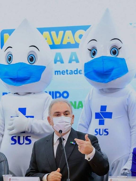 Imagem em primeiro plano mostra o ministro da Saúde, Marcelo Queiroga, de máscara e roupa social sentado de frente para um microfone. Atrás dele, em pé, estão duas pessoas vestidas de Zé Gotinha