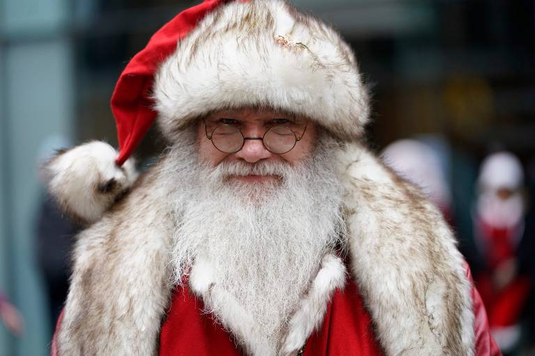 Homem branco e idoso sorri para a câmera. Ele está vestido como o Papai Noel, com gorro de pelos brancos e vermelho. Ele tem uma barba longa branca