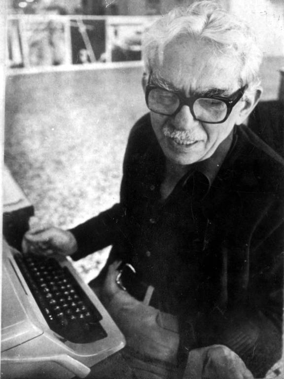 retrato em preto e branco de homem branco idoso, com cabelo e bigodes brancos, vestindo terno e sentado diante de uma máquina de escrever 