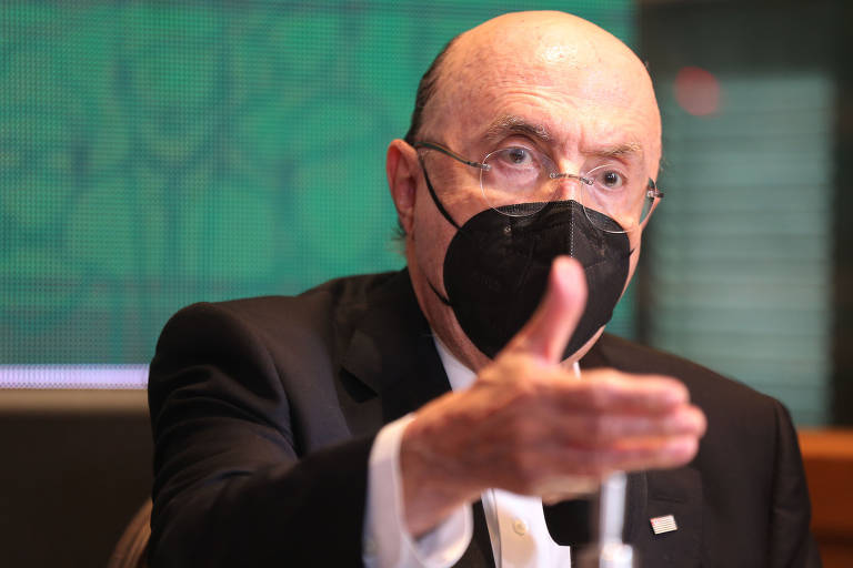 Imagem mostra Henrique Meirelles gesticulando com a mão enquanto fala. Ele usa terno e máscara.