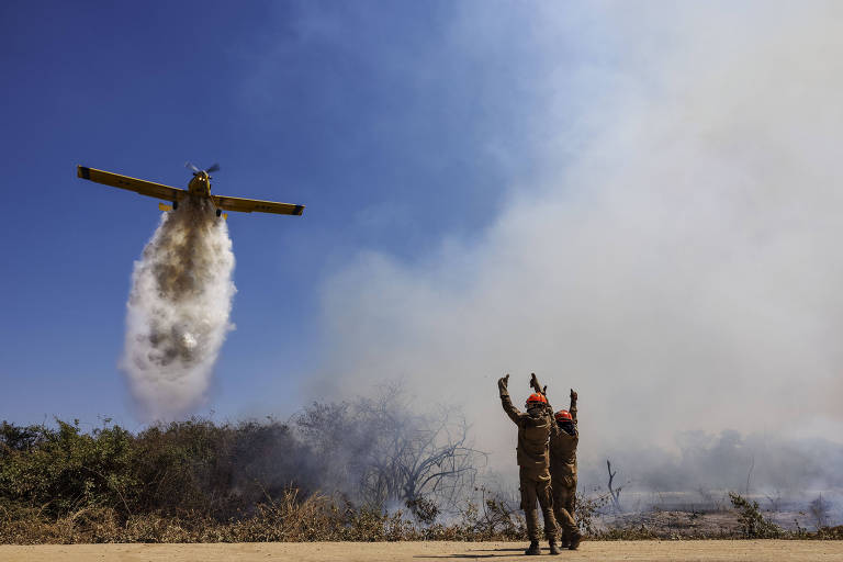 Imagem geram mostra avião descarregando água em meio a fumaça; homens acenam para o avião 