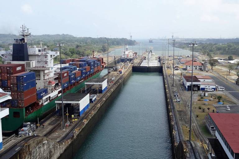Foto mostra o Canal do Panamá visto de cima. É possível ver um corredor de água no centro da foto. À esquerda do corredor, um grande navio de transporte carrega contâiners de diferentes cores
