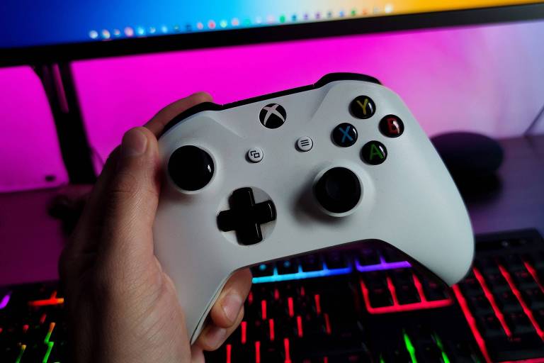 Foto mostra um controle branco do videogame Xbox sendo segurando pela mão de uma pessoa branca. Ao fundo, há um computador com luzes cores rosa e verde, além de uma parede com iluminação neon azul e laranja