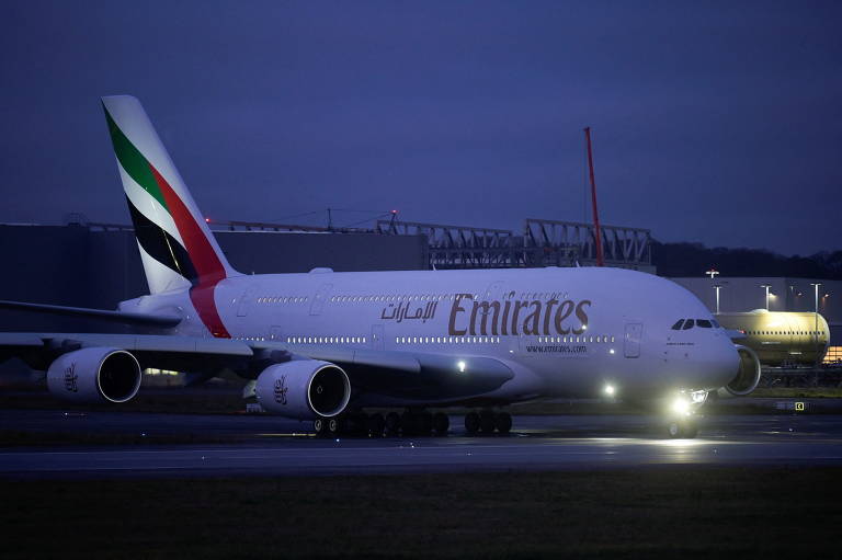 Avião branco, com a inscrição Emirates em dourado e listras verde, vermelha e preta na cauda, pronto para decolar em uma pista de pouso, à noite