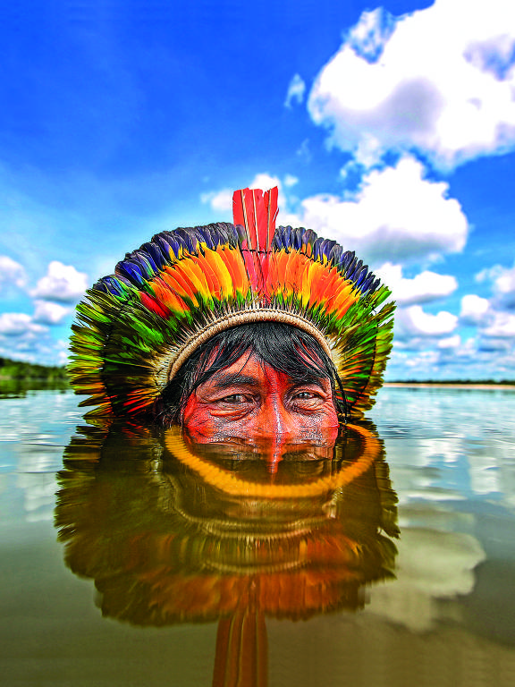 homem indígena usa cocar e afunda metade do rosto no rio