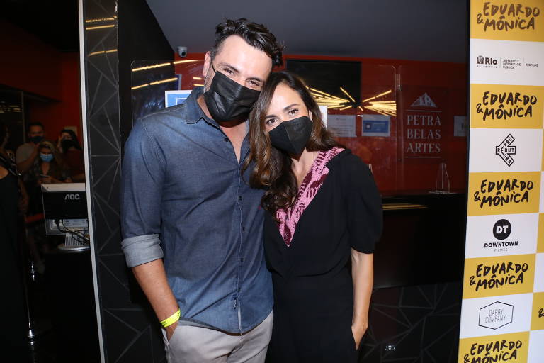 Os atores Rodrigo Lombardi e Alice Braga estão em pé, abraçados; ele us camisa jeans, calça cinza e máscara facial; ela, camisa preta, lenço rosa no pescoço e máscara facial