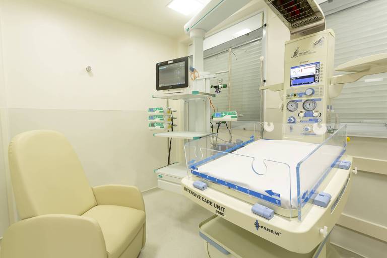 A imagem mostra um quarto de UTI de um hospital, com uma maca coberta por lençóis descartáveis, equipamentos médicos e um monitor na parede. Há uma poltrona no canto inferior esquerdo da imagem para acompanhantes.
