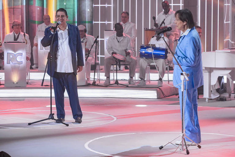 O cantor Zeca Pagodinho está em pé no palco, do lado esquerdo, segurando o microfone; ele usa terno azul marinho. Do lado direito está Roberto Carlos, de terno azul claro, na frente do microfone; ao fundo há uma banda