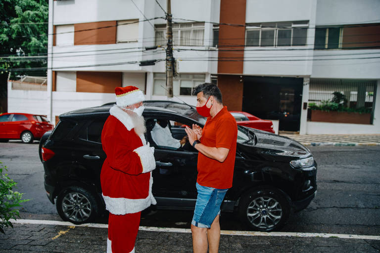 Dois homems em pé, um deles vestido de Papai Noel, conversam frente à frente
