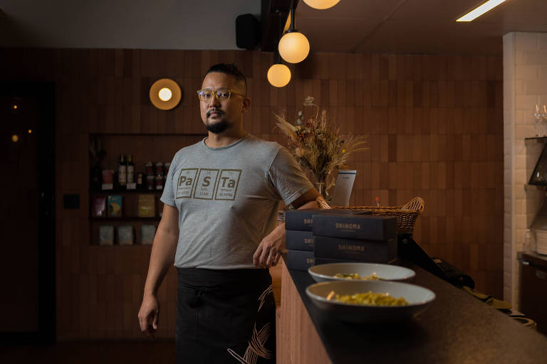 Homem japonês que usa camiseta cinza posa para foto com braço apoiado em uma bancada; nela estão, à direita, pratos  