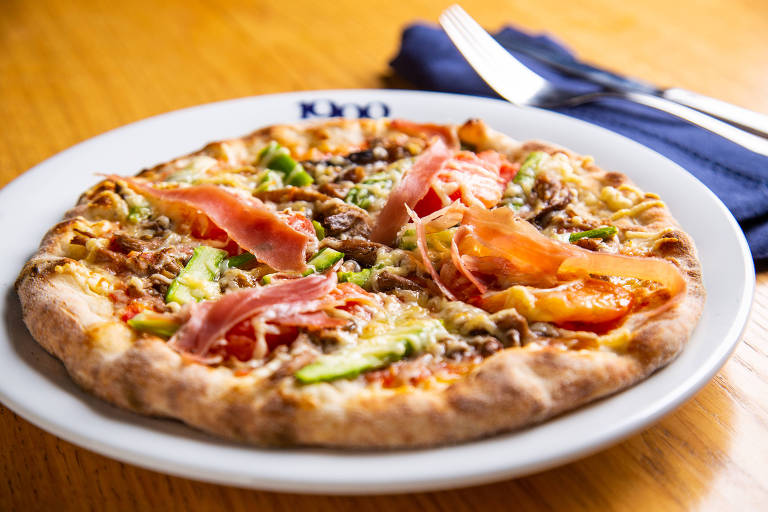 Prato com uma pizza individual em cima de bancada de madeira; à direita, guardanapo azul e talheres  