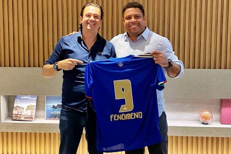 homem de camisa azul escuro segura camisa oficial do Cruzeiro ao lado do jogador Ronaldo Fenômeno