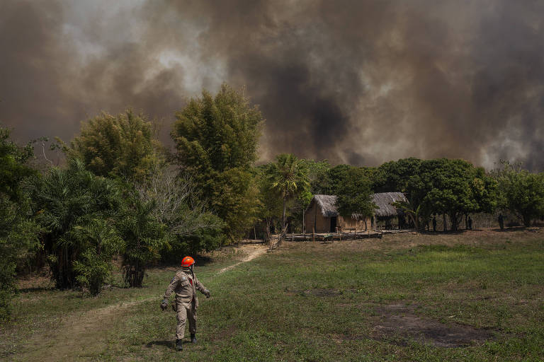 Homem com capacete laranja olha para uma pequena casa, ao fundo há muita fumaça do incêndio