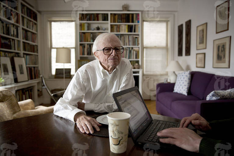 Foto mostra um homem idoso, com cabelos totalmente brancos e óculos, usando uma camisa branca; ele está sentado a uma mesa, numa sala que tem uma estante ao fundo, ladeada por duas janelas, e, ao seu lado, outra pessoa digita num laptop, mas somente suas mãos aparecem na imagem