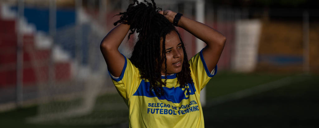 Jovem negra segura os cabelos trançados dentro do campo de futebol, durante treino em que usa uniforme amarelo da seleção feminina de Vitória da Conquista