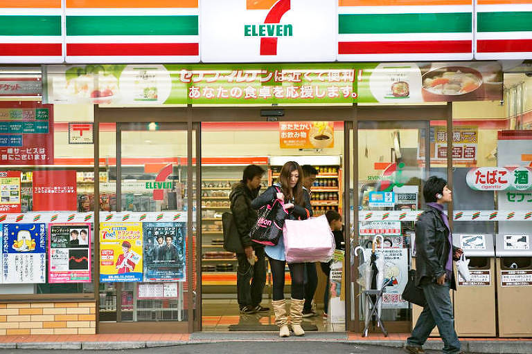 A porta de entrada de uma konbini, no Japão, com vários produtos coloridos e placas de anúncios. Pessoas carregam sacolas saindo da loja, que tem letreiros em rosa, verde e vermelho