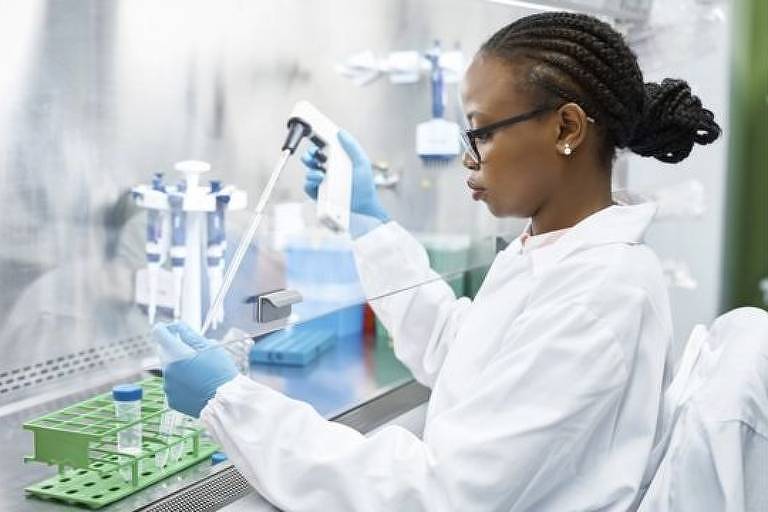 Uma mulher negra está em um laboratório, paramentada com equipamentos de segurança, como jaleco, óculos e luva