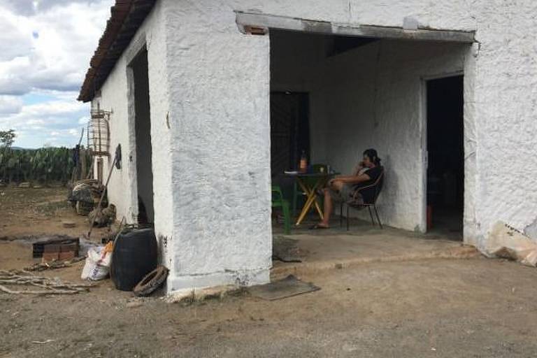 Imagem mostra uma casa em uma área rural. Um homem está sentado em uma cadeira