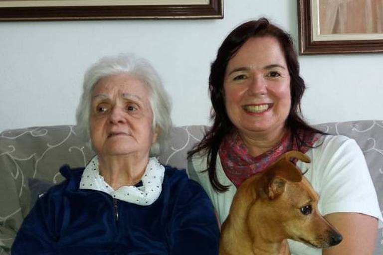 Imagem em primeiro plano mostra mulher idosa ao lado de outra mulher e um cachorro. Elas estão sentadas em um sofá
