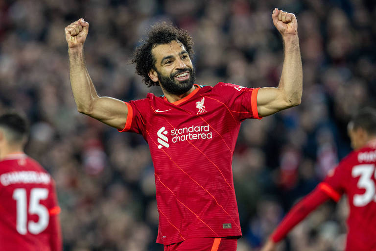 O jogador Mohamed Salah sorri e comemora gol com os dois braços levantados; ele veste o uniforme vermelho do Liverpool