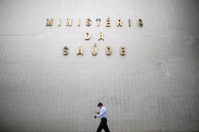 Foto mostra homem com celular na mão passando diante de parede com o letreiro Ministério da Saúde