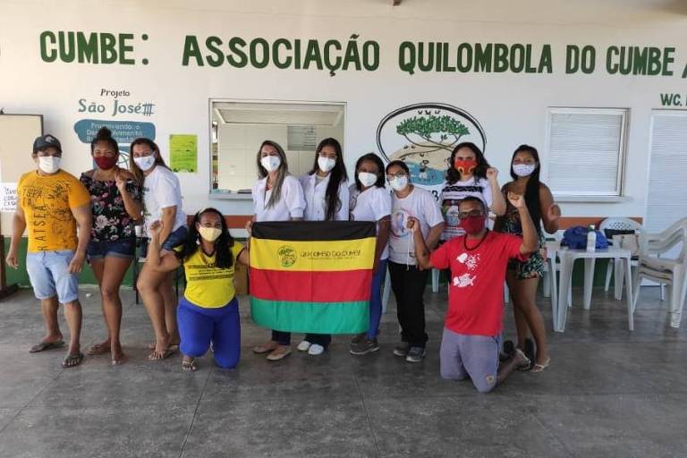 Onze pessoas em pé, lado a lado, seguram em frente a uma parede branca com a inscrição: Associação quilombola do Cumbe