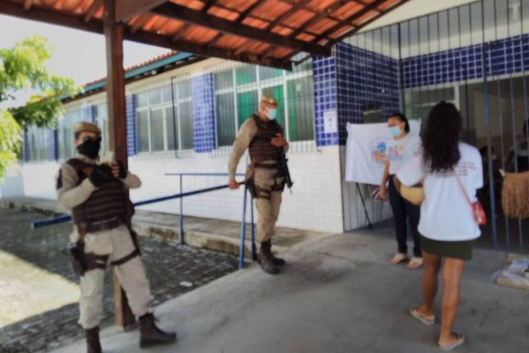 Foto mostra entrada no posto de saúde, dois policiais e duas mulheres aparecem em pé na imagem