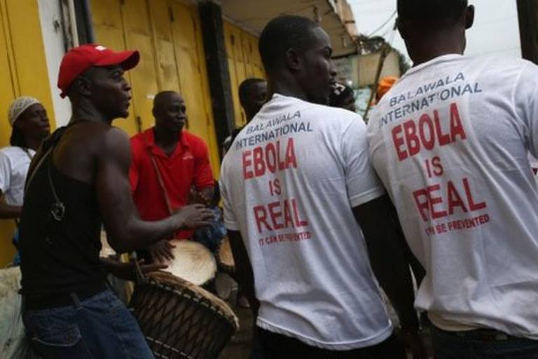Imagem em primeiro plano mostrs homens negros de costas. Dois deles usam camiseta branca escrita: ebola is real