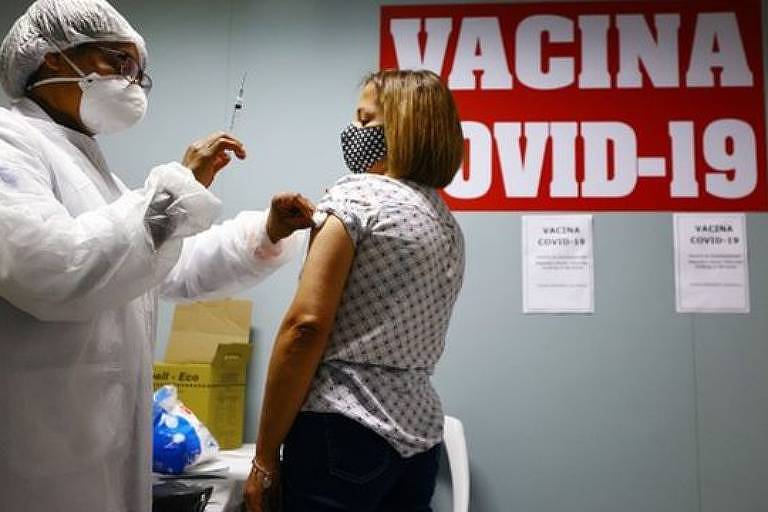 Imagem em primeiro plano mostra profissional da saúde preparando para aplicar vacina no braço de uma mulher.